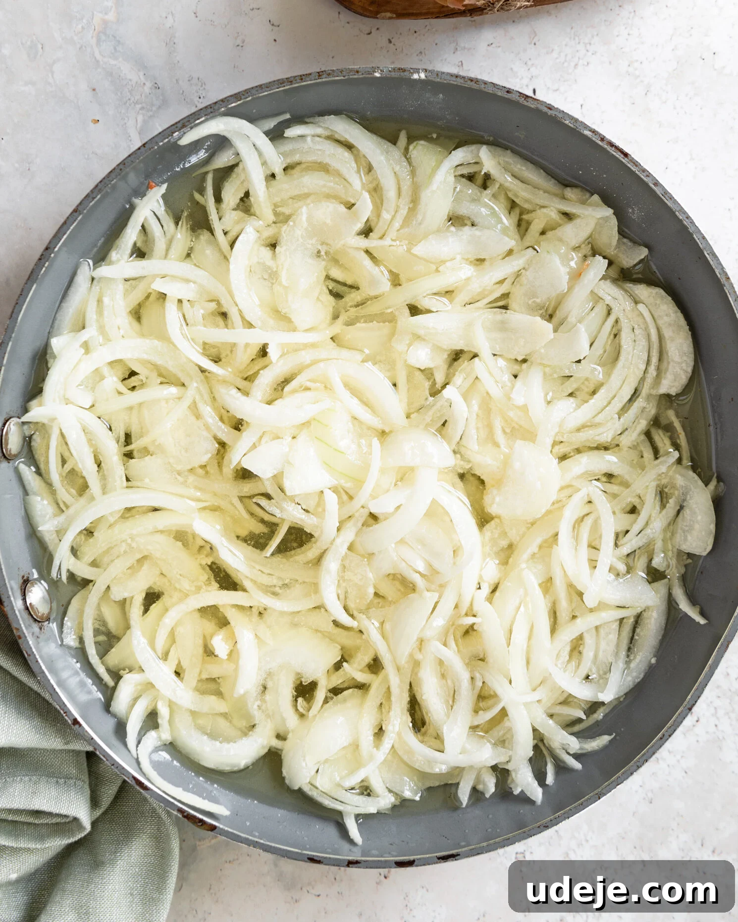 Egypt's Koshari: A Hearty Blend of Rice, Lentils and Pasta 6 sliced white onions in a frying pan with oil with a green towel on a light grey marbled background