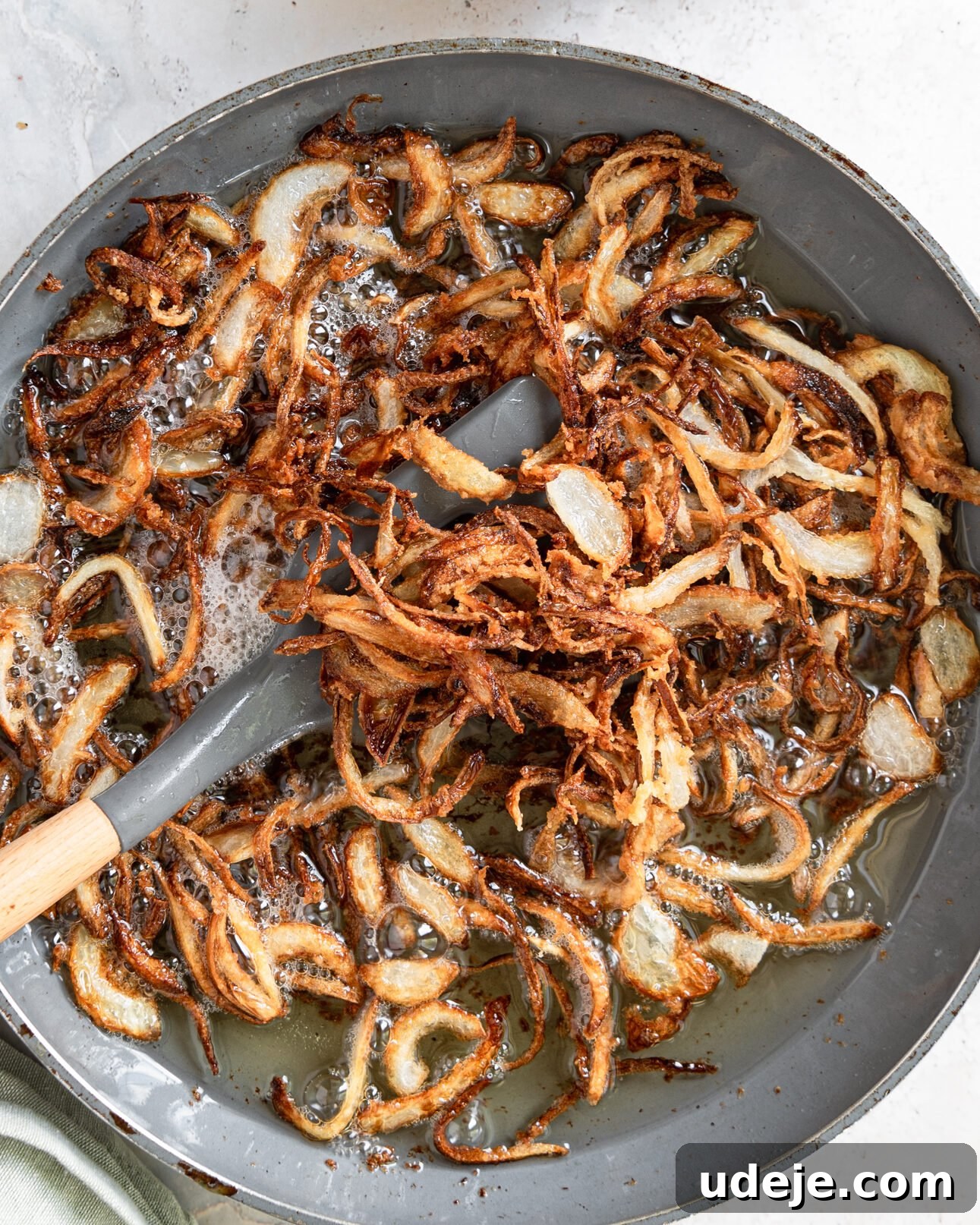 Egypt's Koshari: A Hearty Blend of Rice, Lentils and Pasta 8 dark golden onions in a frying pan with oil and a grey spatula with a green towel on a light grey marble background
