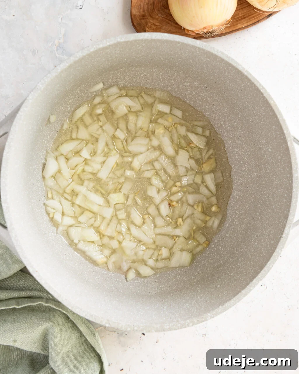 Egypt's Koshari: A Hearty Blend of Rice, Lentils and Pasta 9 chopped onions in a light coloured pot with oil with a green towel on a light grey marble background with a cutting board with onions at the top