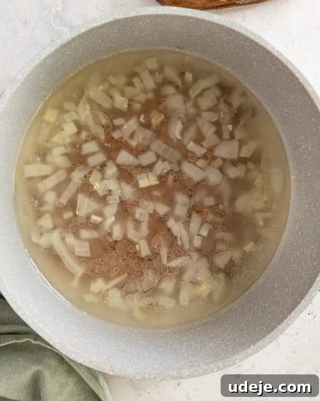 Egypt's Koshari: A Hearty Blend of Rice, Lentils and Pasta 10 chopped onions with brown lentils and broth in a light coloured pot with a green towel on a light grey marble background