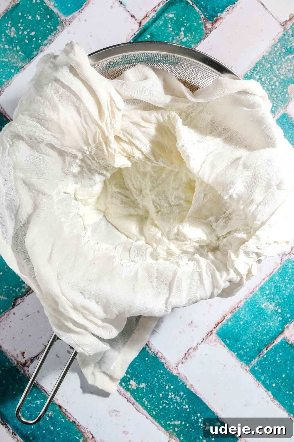 Ali Nazik Smoky Eggplant and Lamb Stew 11 Strained yogurt in an open cheese cloth over a metal mesh strainer over a white bowl on a teal and white brick background