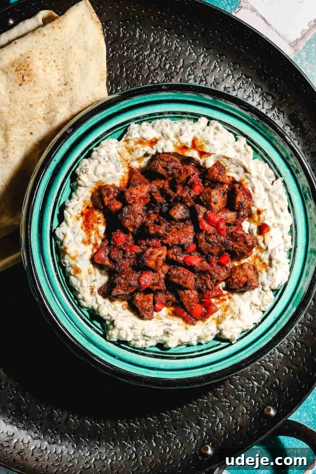 Ali Nazik Smoky Eggplant and Lamb Stew 17 Eggplant and yogurt mixture in a teal and black bowl topped with cooked lamb or veal and red peppers on a black round tray with flat bread to the side on a teal and white background