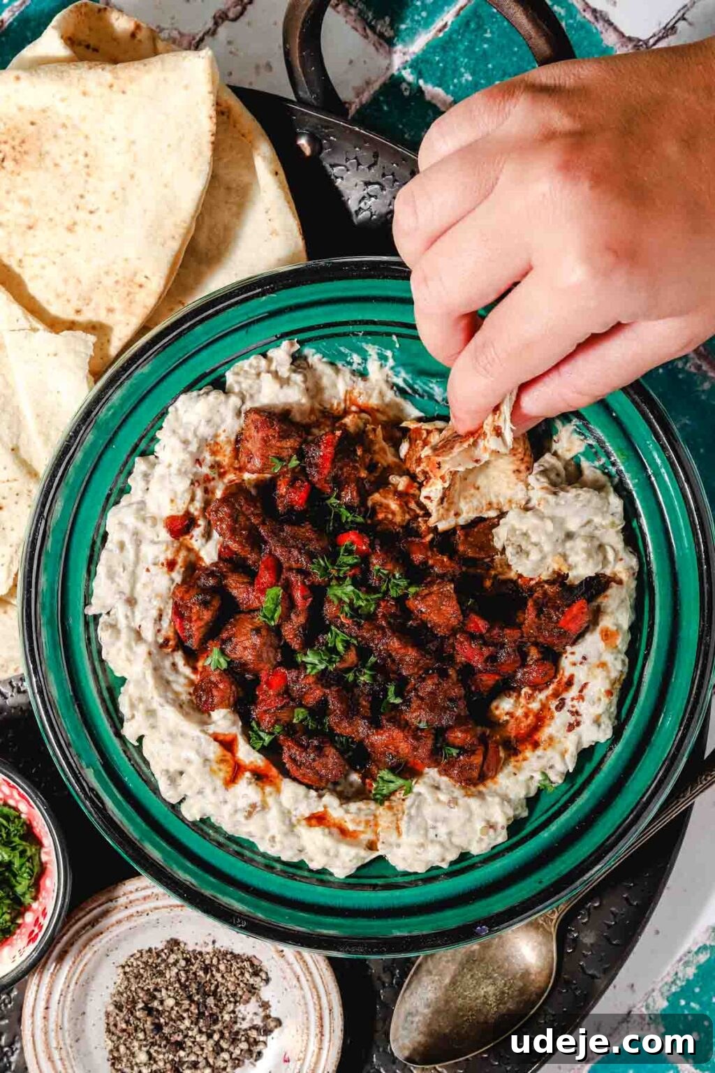 Ali Nazik Smoky Eggplant and Lamb Stew 18 Eggplant and yogurt mixture in a teal and black bowl topped with cooked lamb or veal and red peppers and parsley with a piece of flatbread held by a hand in it on a black round tray with flat bread to the side, a metal spon to the side and small bowls with cracked pepper and parsley on a teal and white background