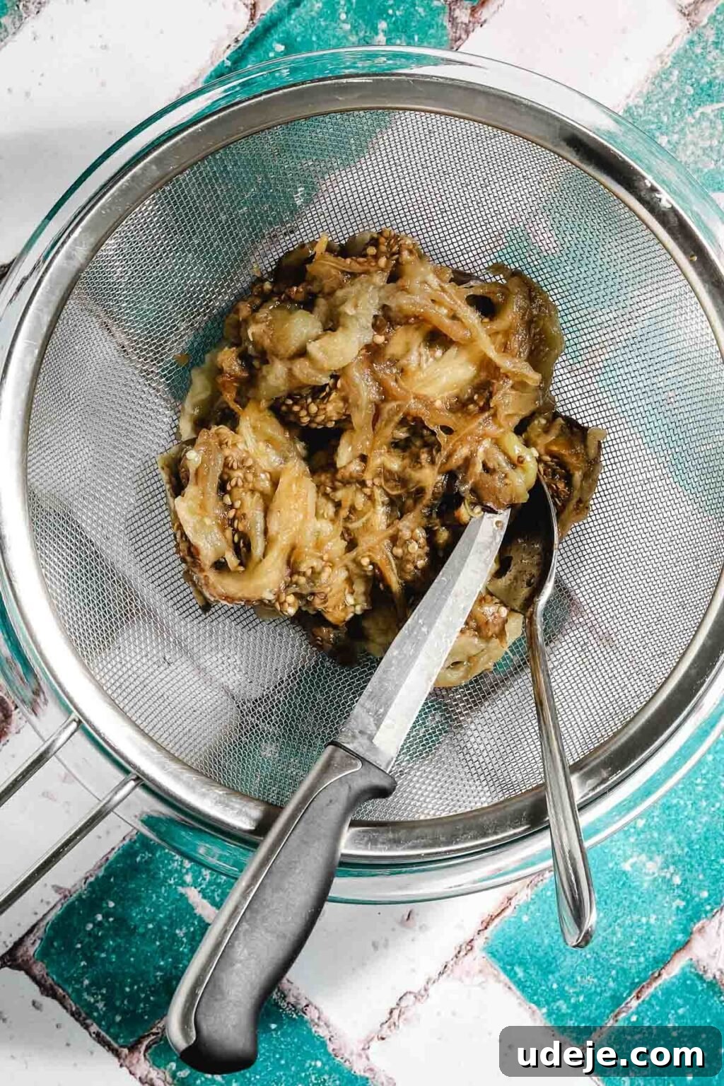 Ali Nazik Smoky Eggplant and Lamb Stew 7 Roasted eggplant flesh in a metal strainer over a glass bowl with a metal spoon and knife in it on a teal and white brick background