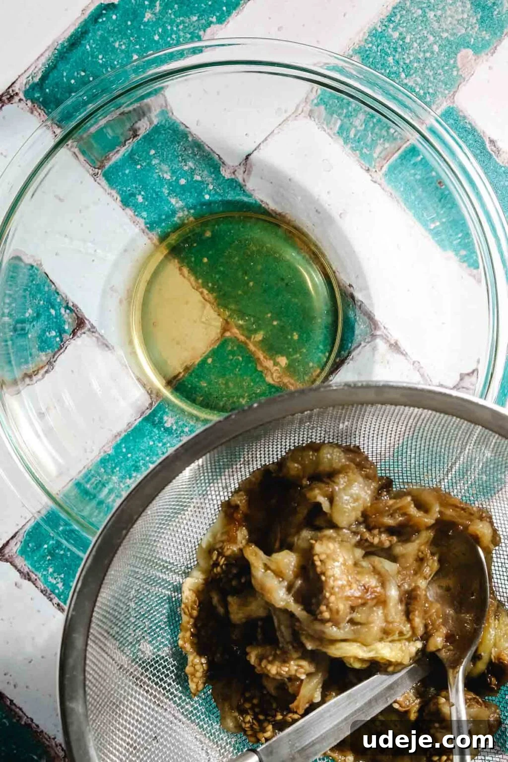 Ali Nazik Smoky Eggplant and Lamb Stew 8 Liquid from strained eggplant flesh in glass bowl with eggplant flesh in a metal mesh strainer on the side of the top of the bowl with a metal spoon and knife in it on a teal and white brick background