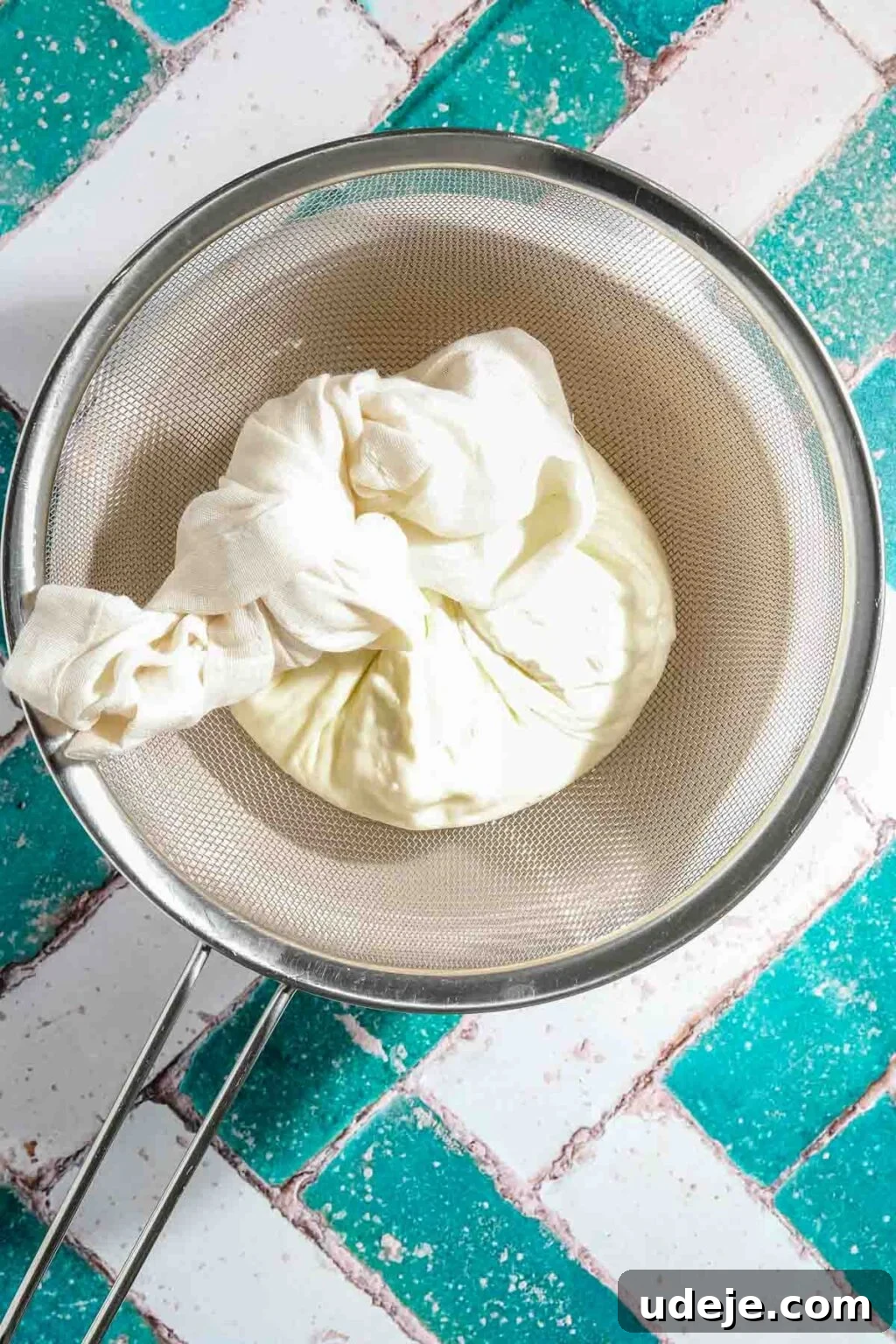Ali Nazik Smoky Eggplant and Lamb Stew 10 Yogurt being strained in a cheese cloth over a metal mesh strainer over a white bowl on a teal and white brick background