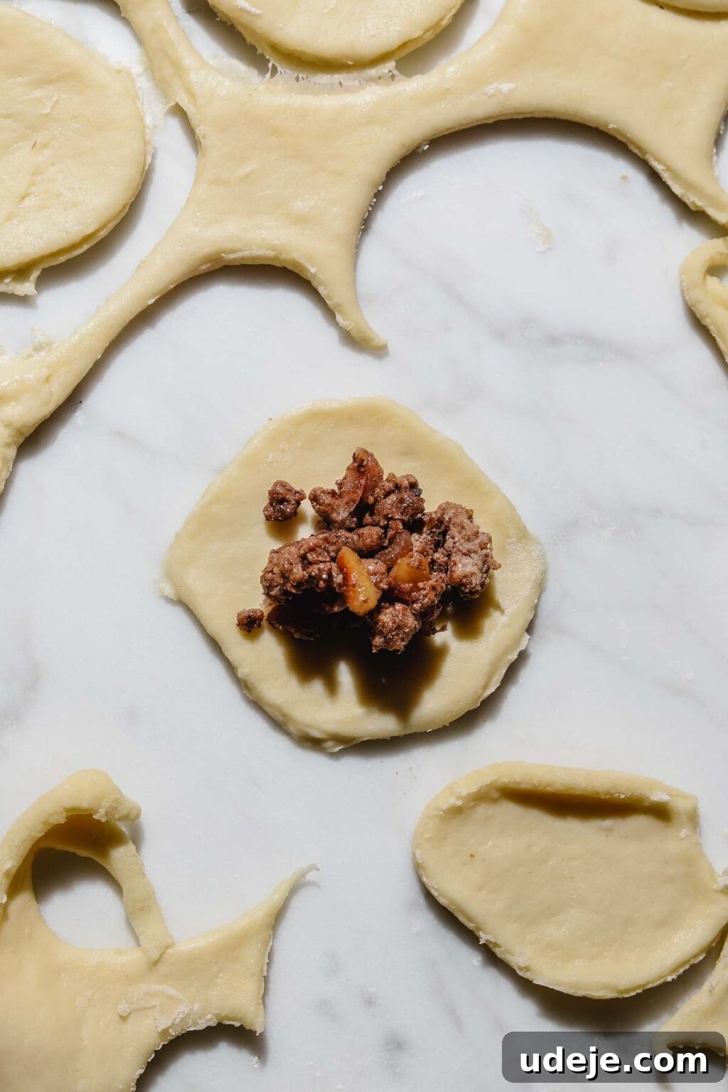 Mastering Homemade Shish Barak: Authentic Yogurt Dumplings 12 A small circular dough cutout with meat filling positioned in the center, surrounded by other dough pieces and scraps on a light marble background.