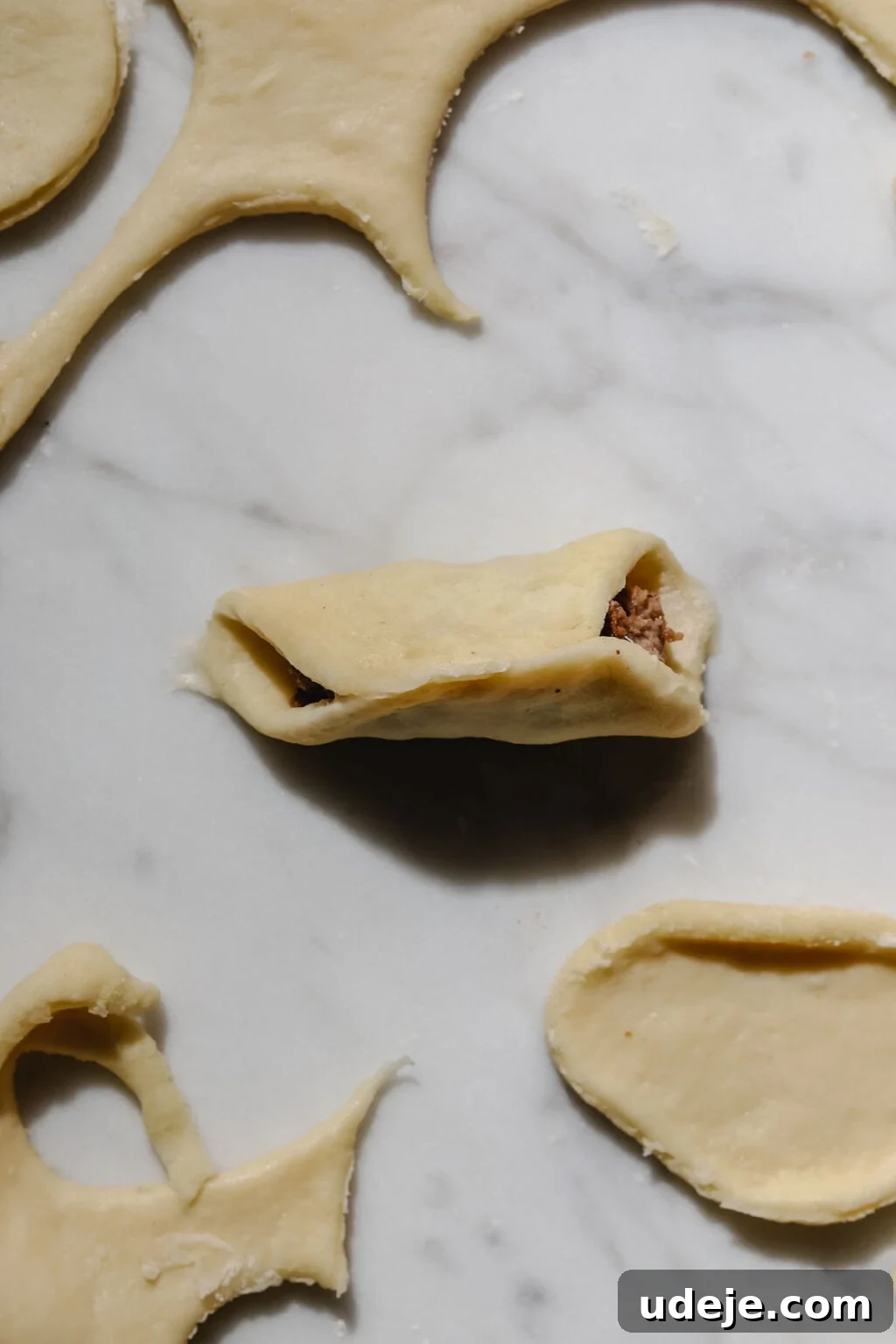 Mastering Homemade Shish Barak: Authentic Yogurt Dumplings 13 A small circular dough cutout, folded over into a half-moon shape with the meat filling enclosed, alongside other dough pieces and scraps on a light marble background.