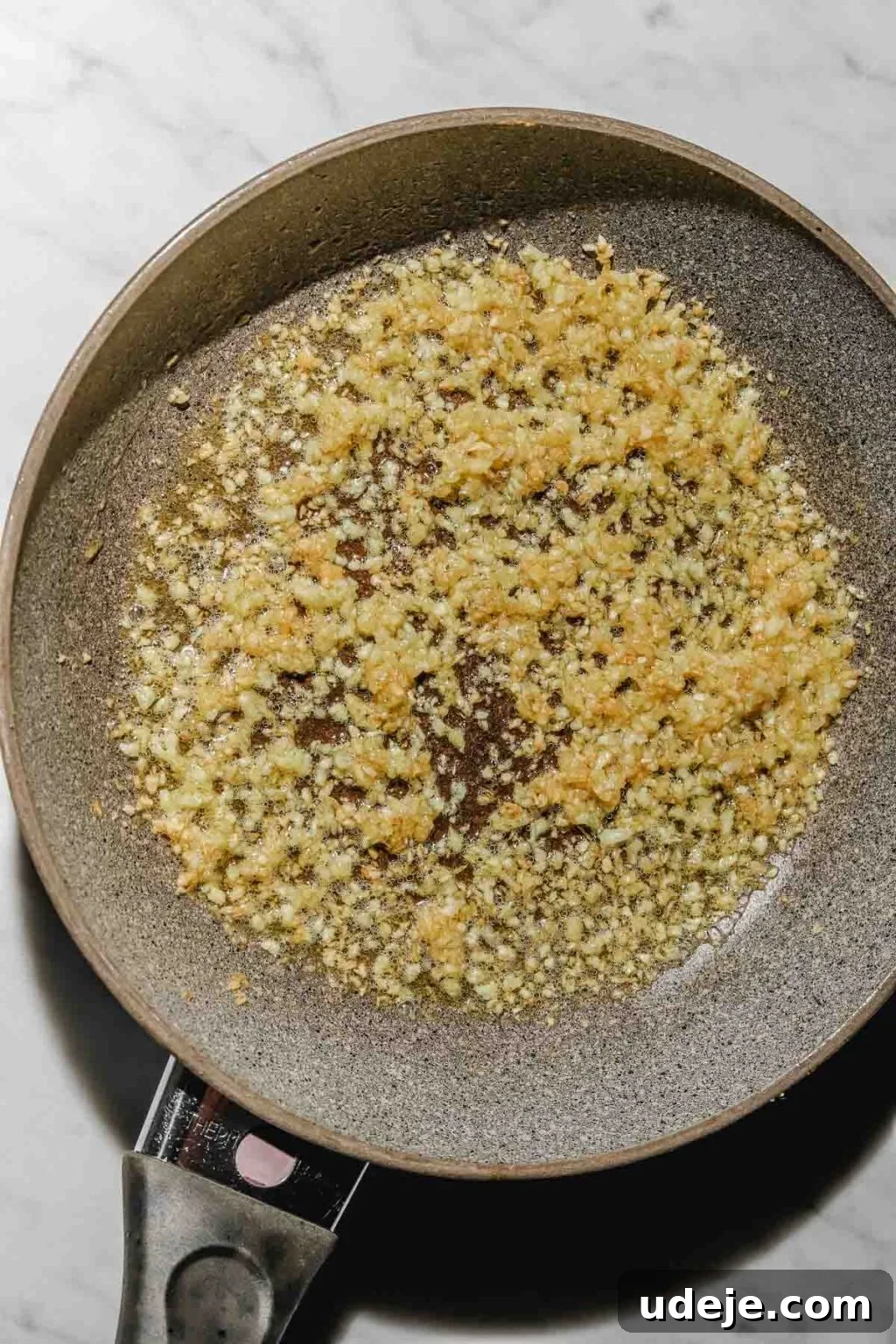 Mastering Homemade Shish Barak: Authentic Yogurt Dumplings 19 Minced garlic cooking in olive oil in a small sauté pan on a light marble background, releasing its fragrant aroma.