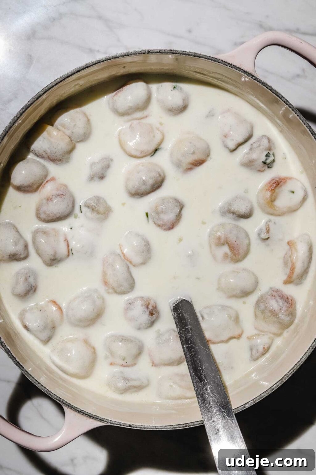 Mastering Homemade Shish Barak: Authentic Yogurt Dumplings 20 Shish Barak dumplings luxuriating in the creamy yogurt sauce in a pink-handled pot, with a metal ladle ready for serving, all on a light marble background.