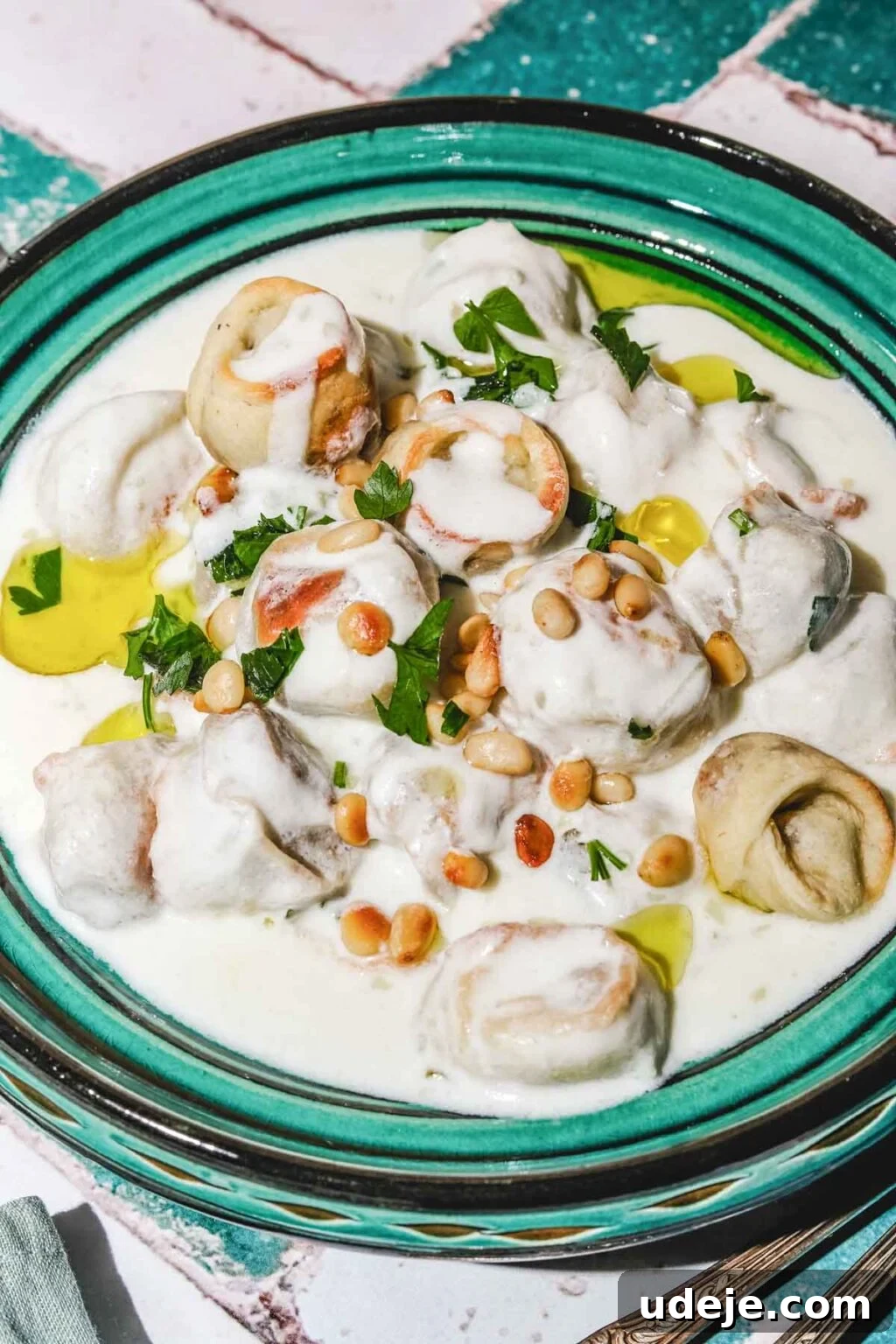 Mastering Homemade Shish Barak: Authentic Yogurt Dumplings 21 Shish Barak, featuring delectable meat-filled dumplings served in a creamy yogurt sauce on a teal-colored plate, beautifully garnished with toasted pine nuts, fresh chopped parsley, and a delicate drizzle of olive oil.
