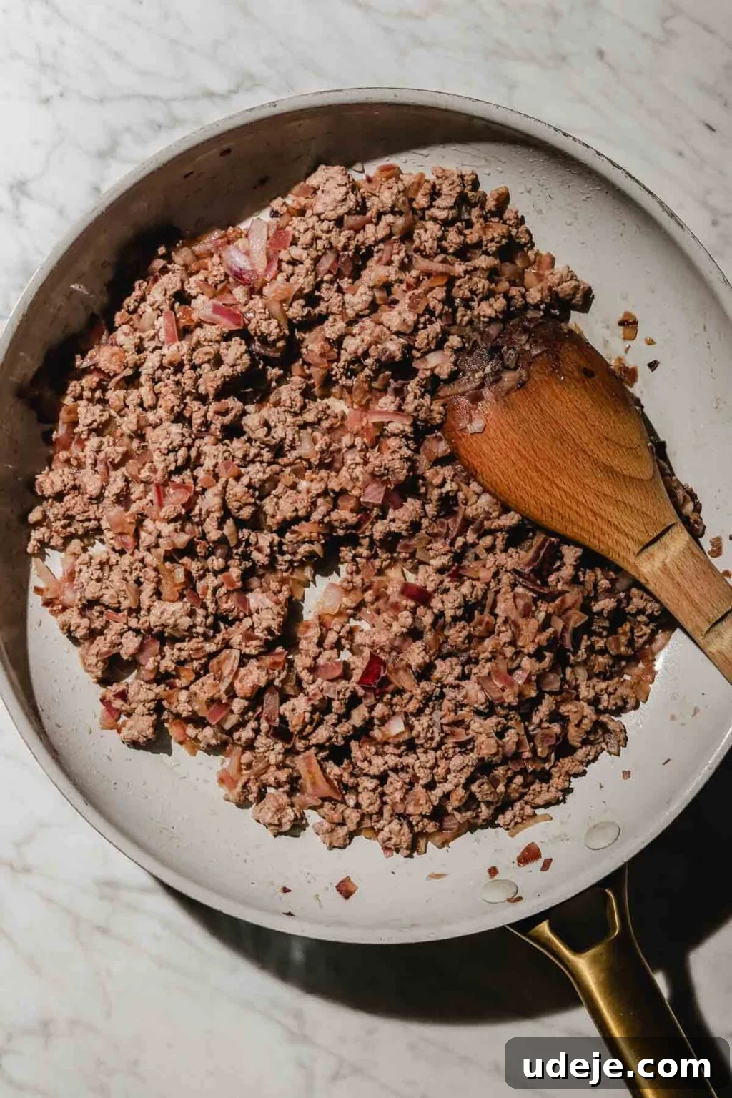 Mastering Homemade Shish Barak: Authentic Yogurt Dumplings 6 The Shish Barak filling in progress, with softened onions and browning ground beef in a light grey skillet, stirred with a wooden spoon on a light marble background.
