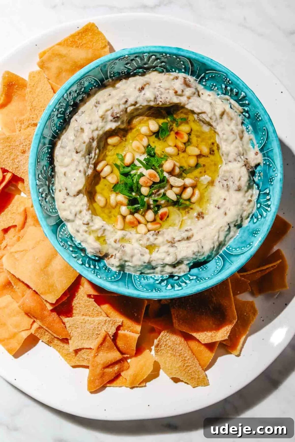 Smoky Mutabal: A Creamy Tahini Treat 2 Mutabal in a blue patterned bowl with olive oil, pine nuts and parsley on a large white plate surrounded by pita chips on a light marble background