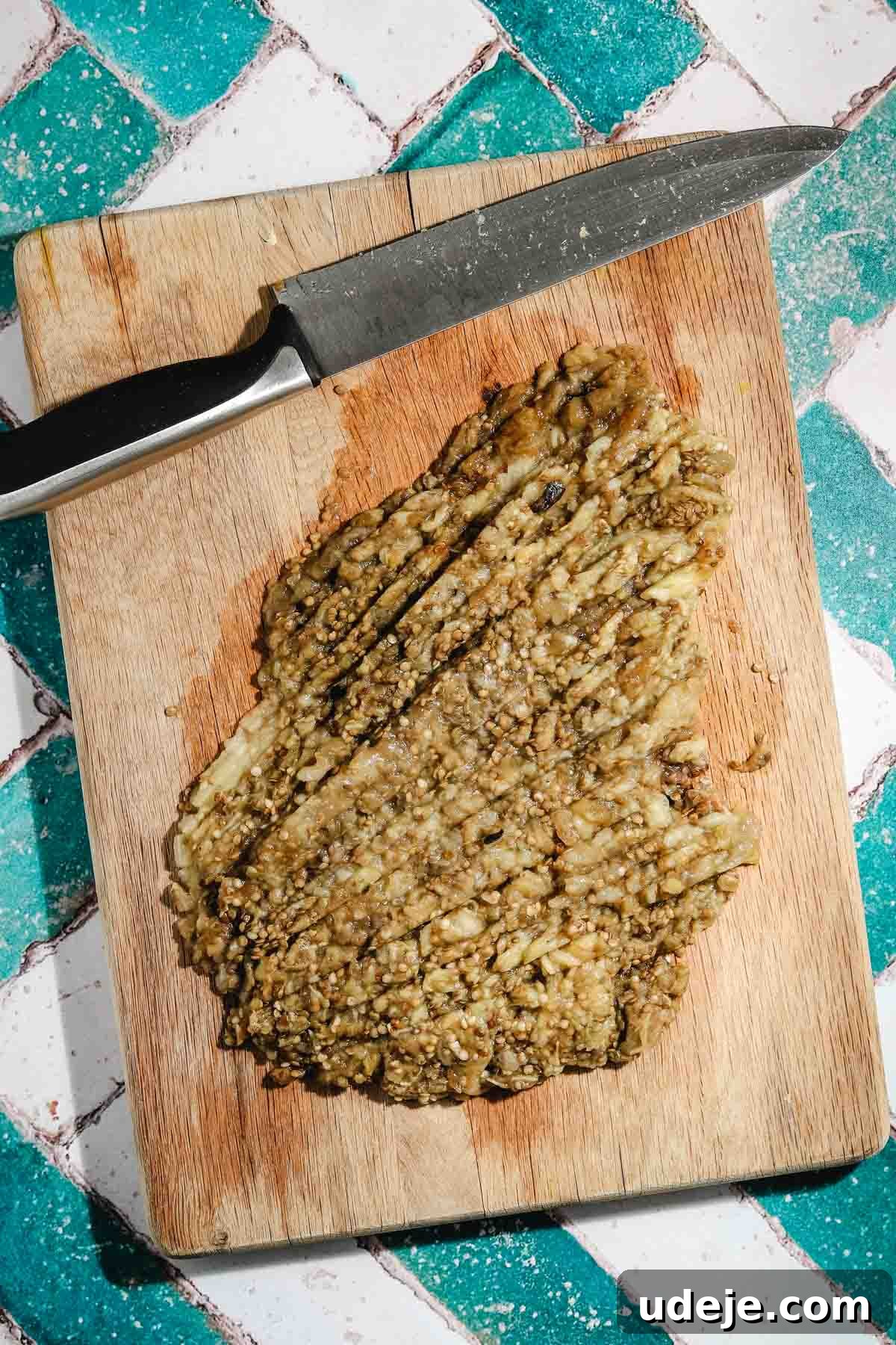 Smoky Mutabal: A Creamy Tahini Treat 11 Chopped roasted and strained eggplant flesh on a wooden cutting board with a large knife to the top on a teal and white brick background