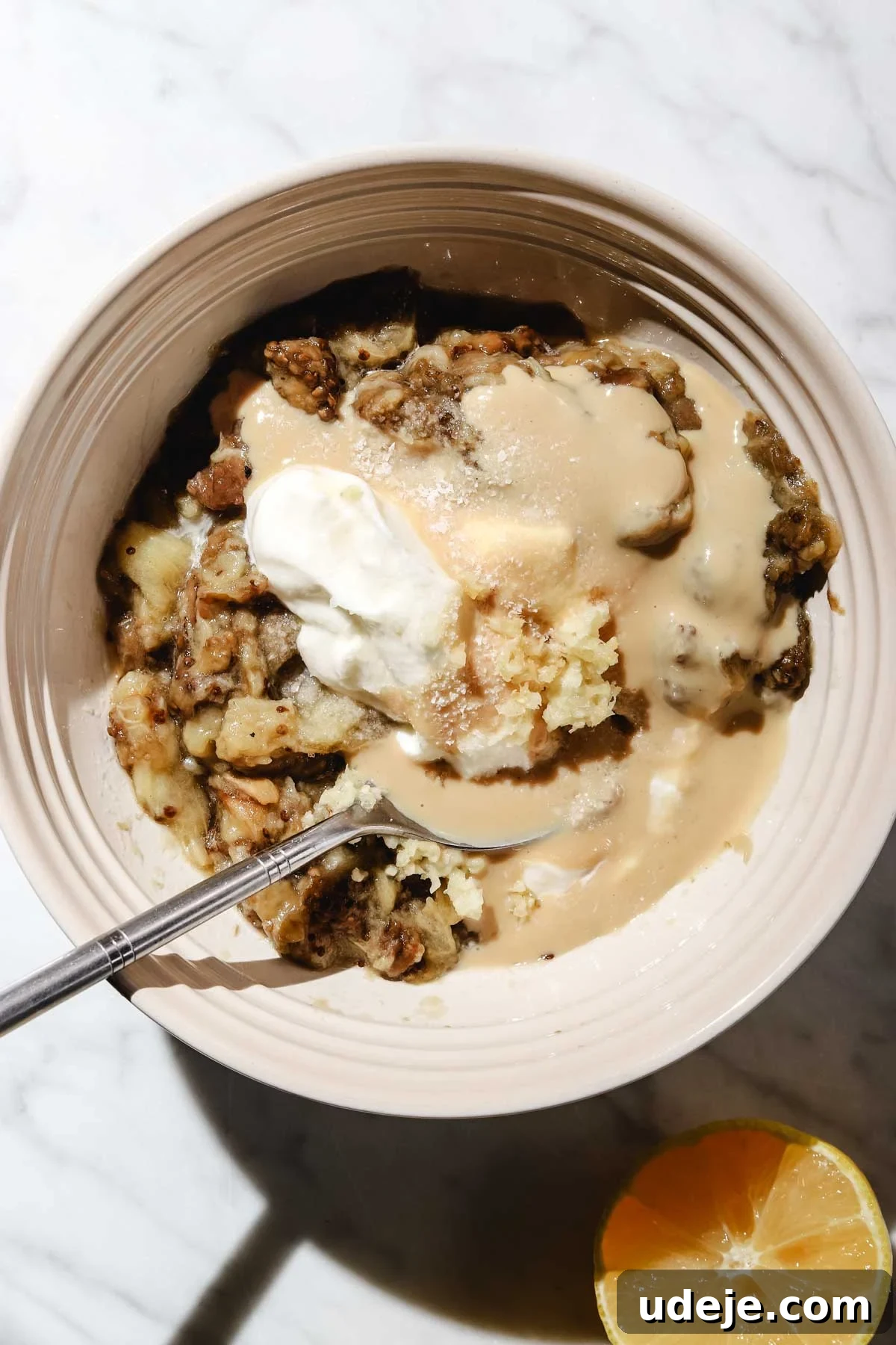 Smoky Mutabal: A Creamy Tahini Treat 12 Mashed eggplant with garlic, yogurt and tahini in a small bowl with a spoon in it with a half lemon beside it on a light marble background