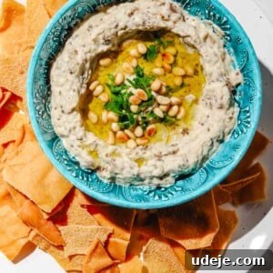 Mutabal in a blue patterned bowl with olive oil, pine nuts and parsley on a large white plate surrounded by pita chips on a light marble background