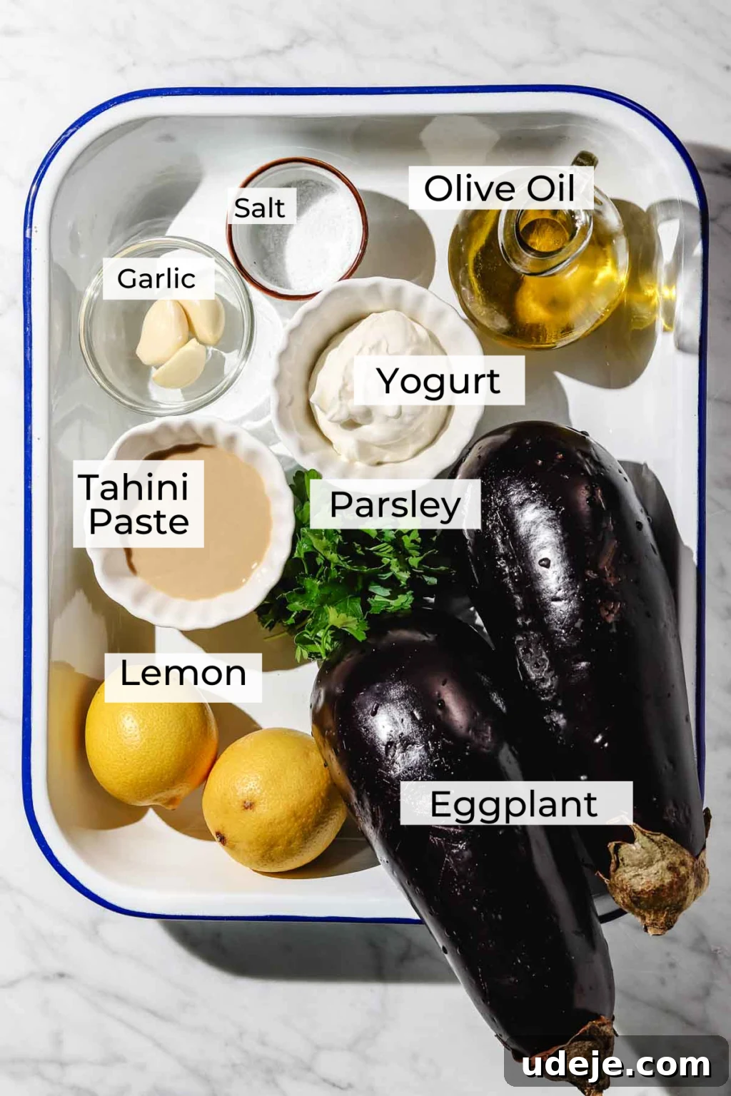 Smoky Mutabal: A Creamy Tahini Treat 4 Mutabal ingredient shot with small pitcher of olive oil, small bowls of salt, garlic cloves, yogurt, tahini paste, small bunch of parsleym 2 lemons and 2 large eggplants in a blue rimmed white baking pan on a light marble background