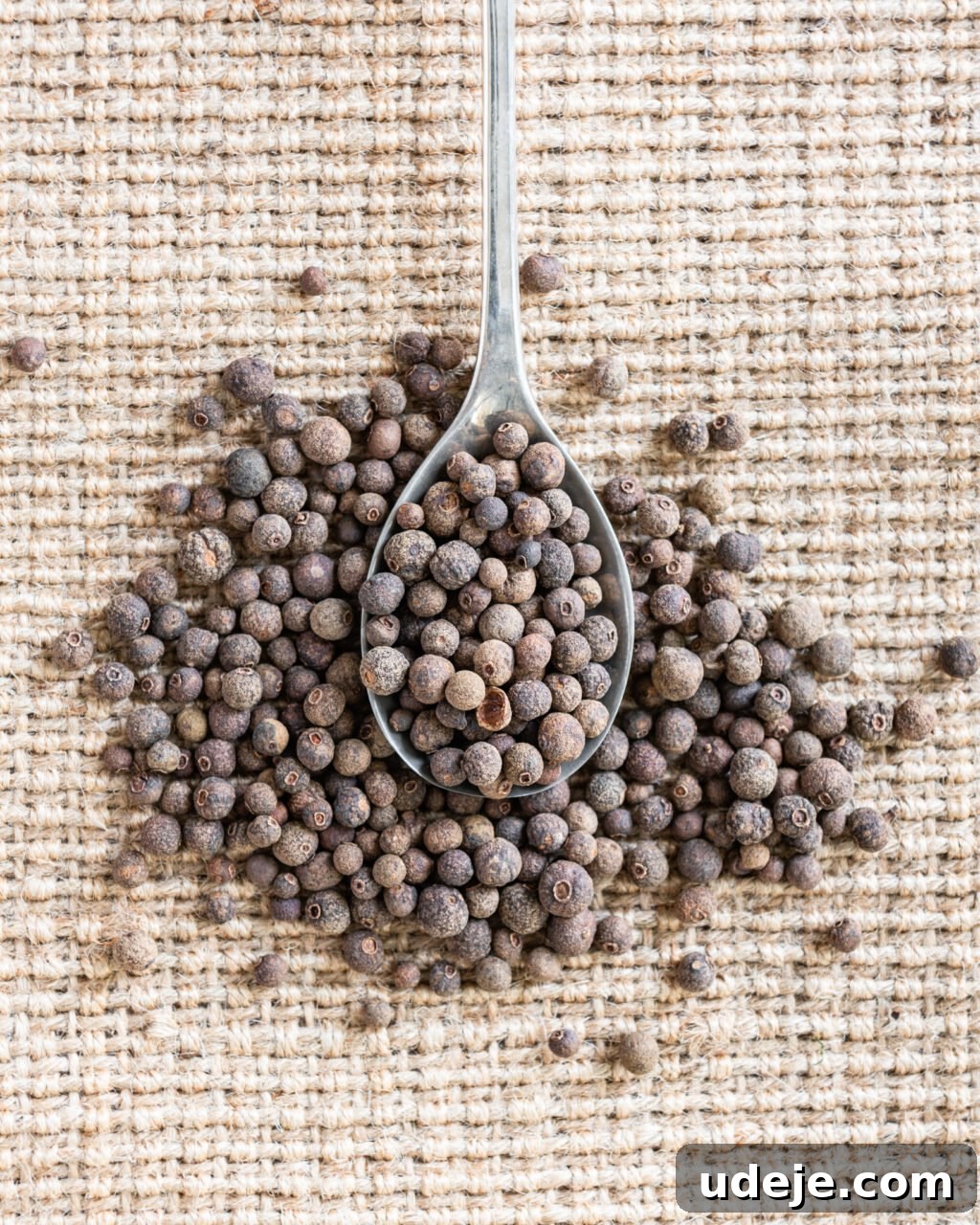 Unlocking the Flavors of Allspice in Middle Eastern Dishes 2 Whole allspice on a metal spoon surrounded by whole allspice on a woven beige background