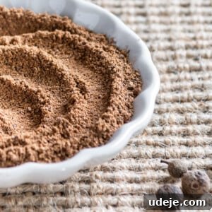 Ground allspice in a small white scalloped dish with whole allspice beside it on a beige woven background