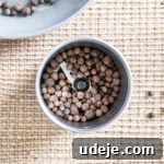 whole allspice in a spice grinder with a light grey skillet in the top corner on a beige woven background