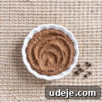 Ground allspice in a small scalloped dish with some whole allspice beside it on a beige woven background