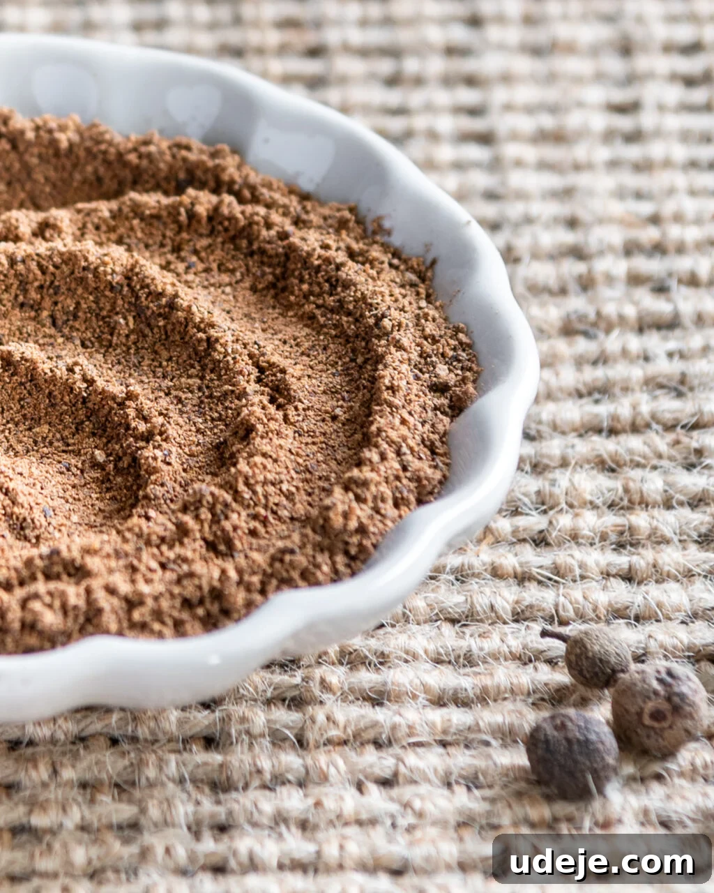 Unlocking the Flavors of Allspice in Middle Eastern Dishes 3 Ground allspice in a small white scalloped dish with whole allspice beside it on a beige woven background