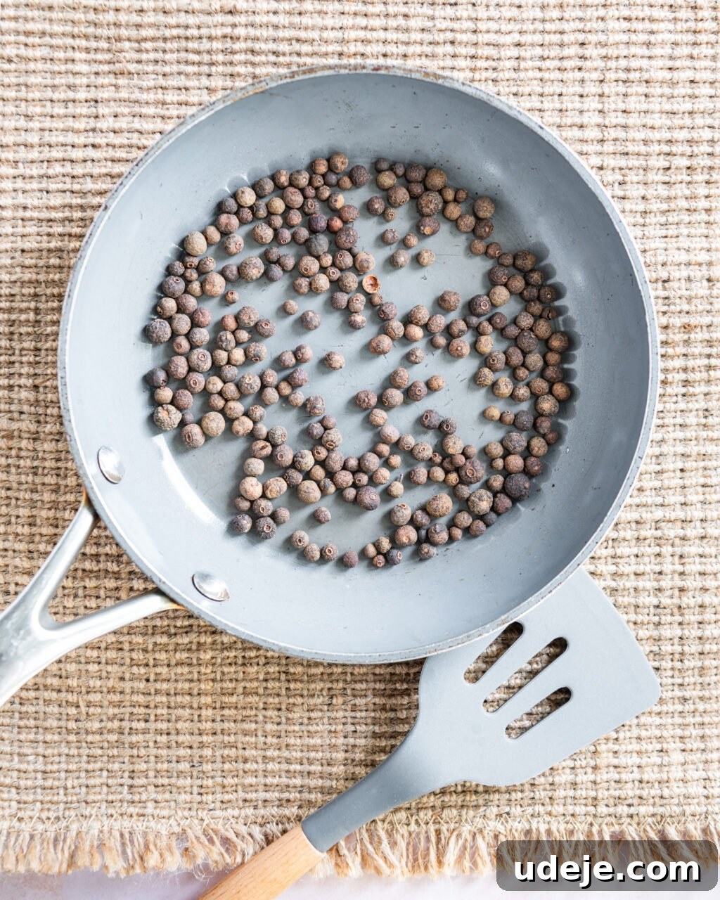 Unlocking the Flavors of Allspice in Middle Eastern Dishes 4 light grey skillet with whole allspice with a light grey spatula beside the skillet on a light beige woven background