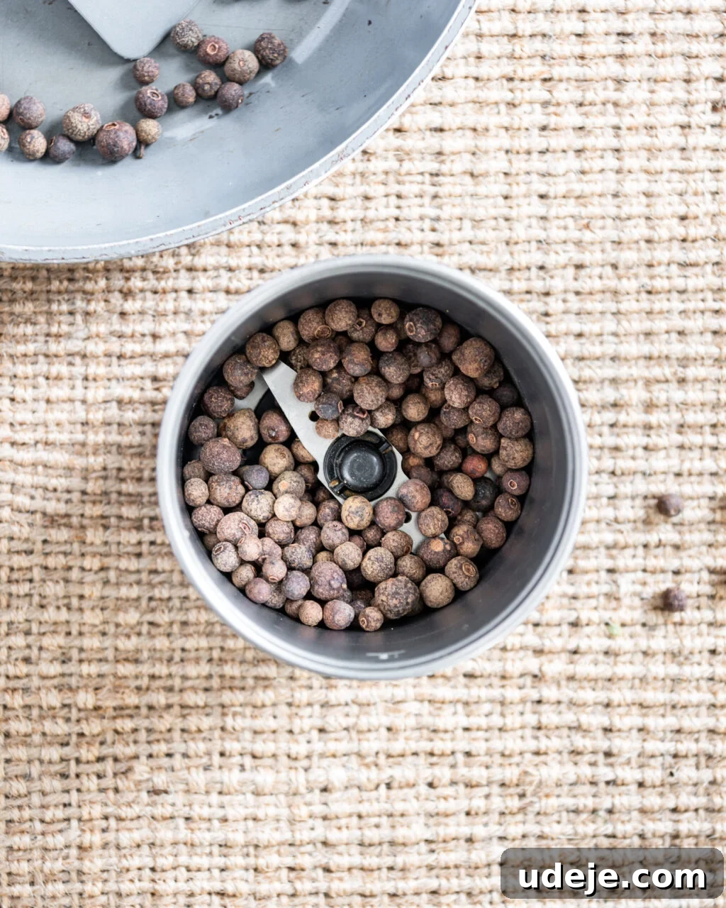 Unlocking the Flavors of Allspice in Middle Eastern Dishes 5 whole allspice in a spice grinder with a light grey skillet in the top corner on a beige woven background