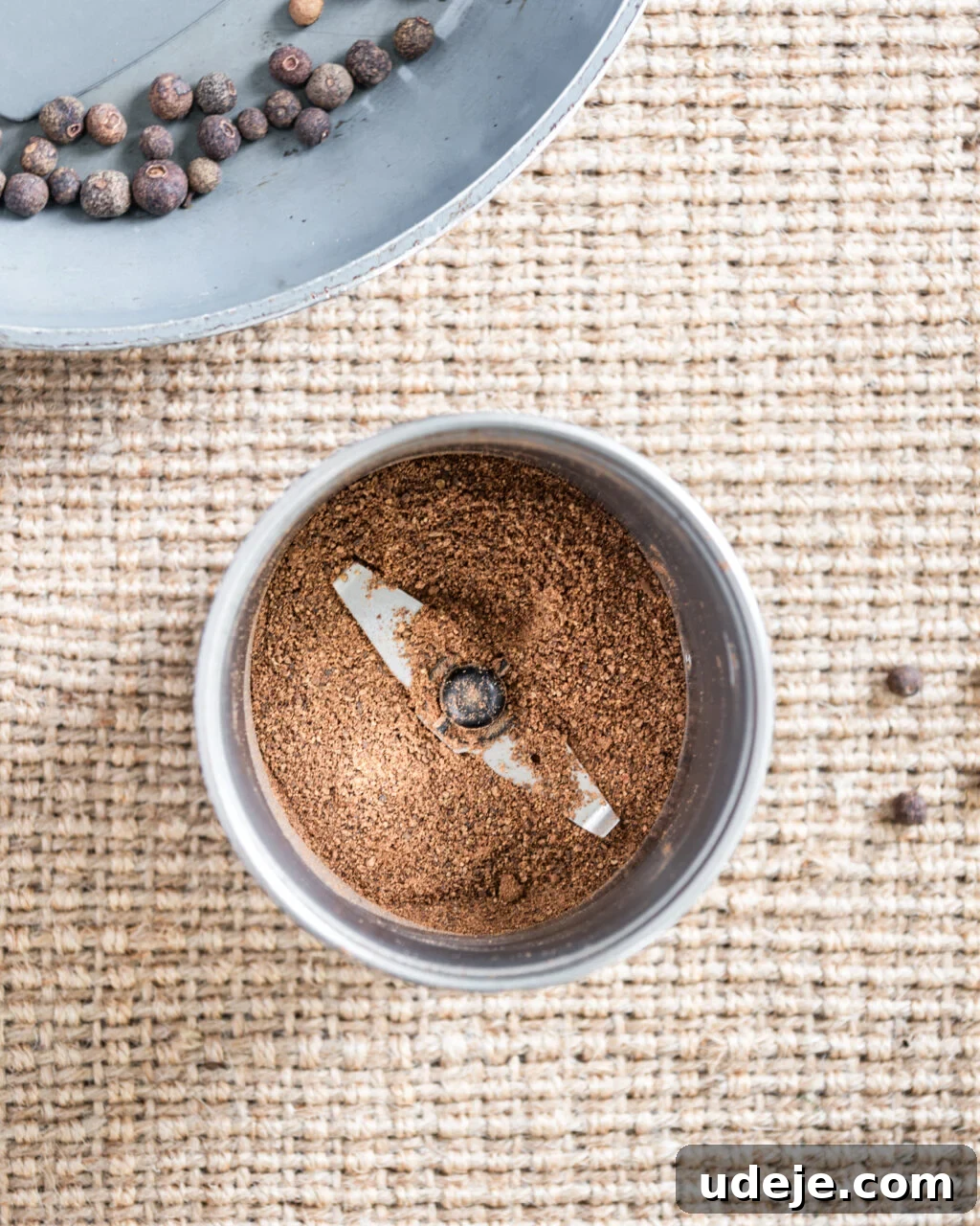 Unlocking the Flavors of Allspice in Middle Eastern Dishes 6 Ground allspice in a spice blender with a light grey skillet with whole allspice in the top corner on a beige woven background