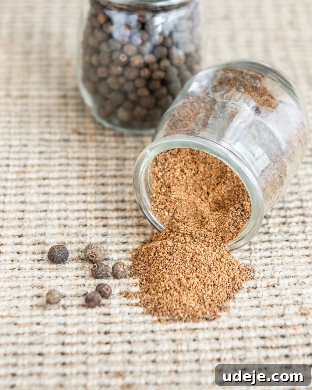 Unlocking the Flavors of Allspice in Middle Eastern Dishes 7 Ground allspice spilling from a small glass jar on it's side with whole allspice in a glass jar behind it and some whole allspice on a beige woven background