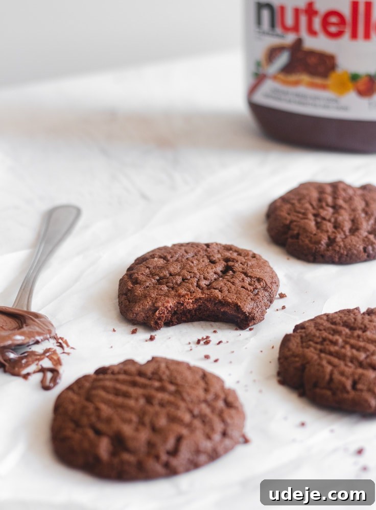 Nutella Cookie Bliss in 15 Minutes 2 Nutella cookies on parchment paper with a spoonful of Nutella next to them and a jar of Nutella to the back