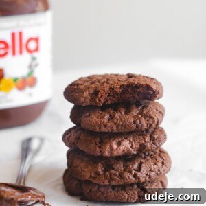 Nutella Cookie Bliss in 15 Minutes 8 A stack of five Nutella Cookies sits on white parchment paper, with a jar of Nutella in the background and a spoonful of chocolate spread in the foreground. One cookie on top has a bite taken out.