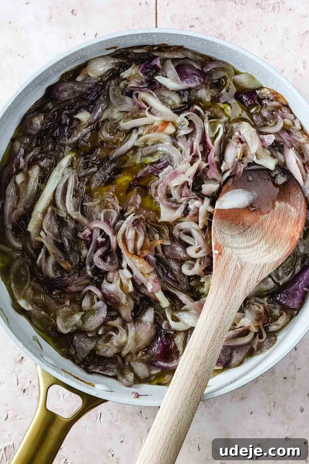 Sumac-Spiced Chicken Musakhan Crisps 11 Caramelized red onions in a light coloured skillet with a gold handle with a wooden spoon on a light coloured tile background, showcasing their golden-brown hue.