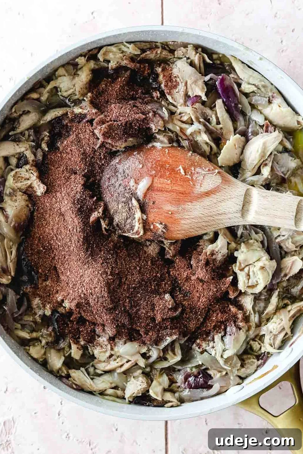 Sumac-Spiced Chicken Musakhan Crisps 12 Shredded chicken in a skillet with spices and a wooden spoon on a light coloured background, ready to be mixed with onions.