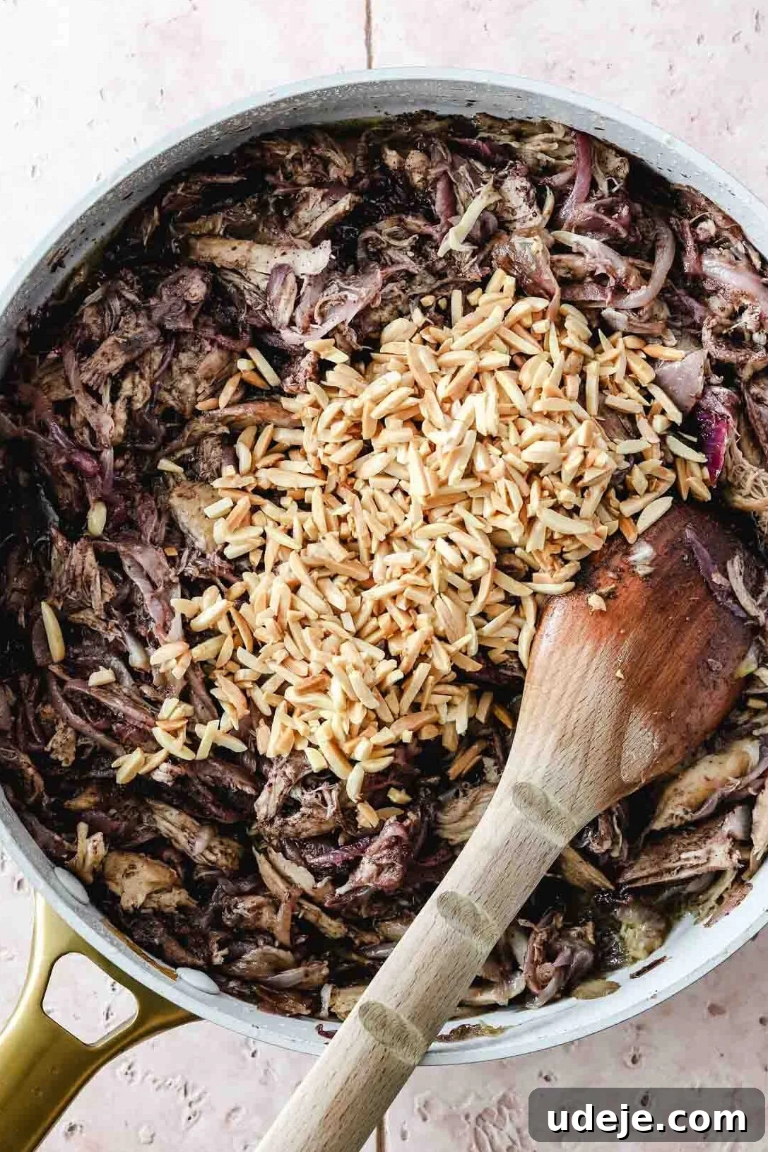Sumac-Spiced Chicken Musakhan Crisps 13 Shredded chicken mixed with caramelized onions and slivered almonds in a light coloured skillet with a wooden spoon on a light coloured tiled background, a fragrant filling.