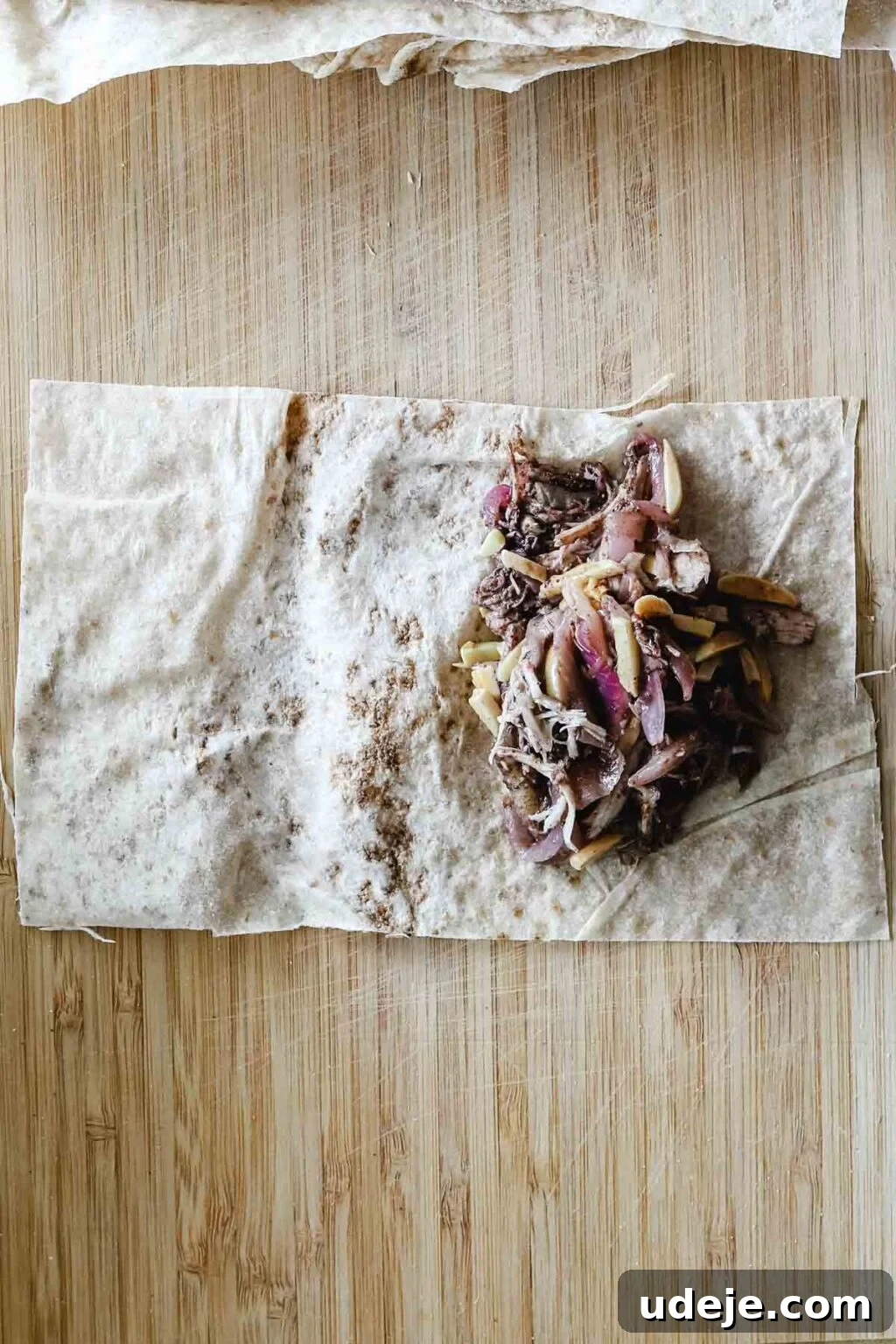 Sumac-Spiced Chicken Musakhan Crisps 15 Rectangular piece of Markouk with shredded chicken and onion filling on a light coloured wooden cutting board, prepped for rolling.