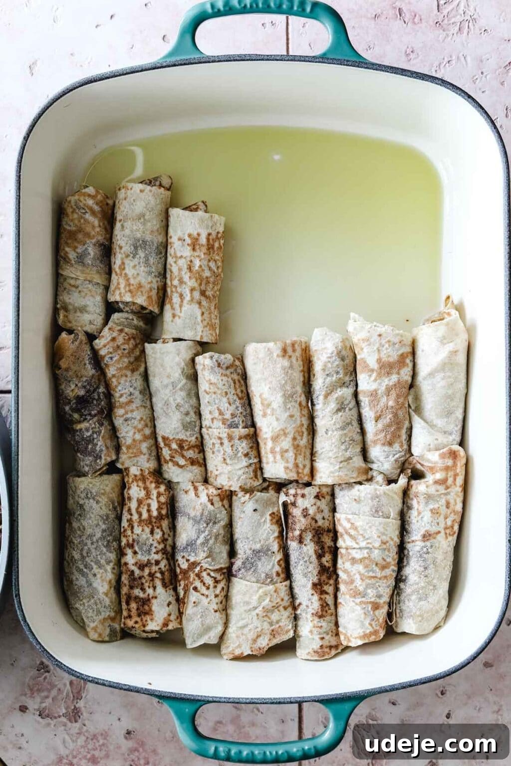 Sumac-Spiced Chicken Musakhan Crisps 16 Musakhan rolls in a half-filled teal and white coloured baking dish on a light coloured tiled background, neatly arranged.