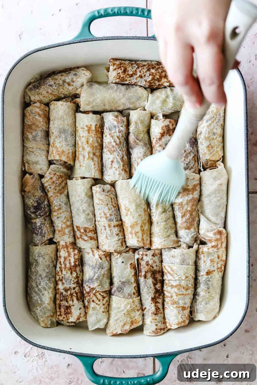 Sumac-Spiced Chicken Musakhan Crisps 17 Musakhan rolls in a teal and white coloured baking dish being brushed with olive oil on a light coloured tiled background, prior to baking.