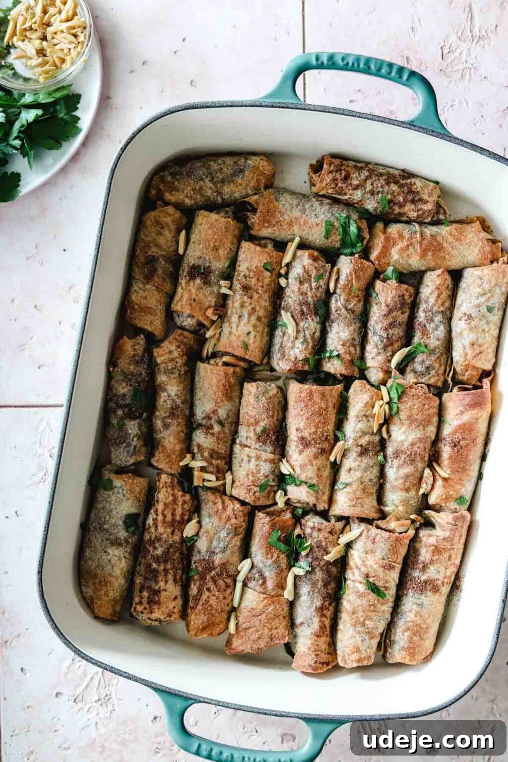 Sumac-Spiced Chicken Musakhan Crisps 18 Baked Musakhan rolls in a teal and white coloured baking dish on a light coloured tiled background with a white plate with parsley and slivered almonds, garnished and ready to serve.