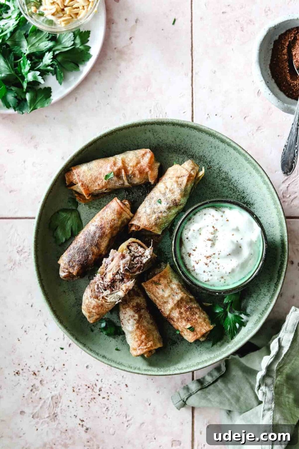 Sumac-Spiced Chicken Musakhan Crisps 19 Musakhan rolls in a green plate with a yogurt dip in a small bowl on a light coloured tile background with a bowl of sumac an a spoon and a light green tea towel, beautifully arranged.