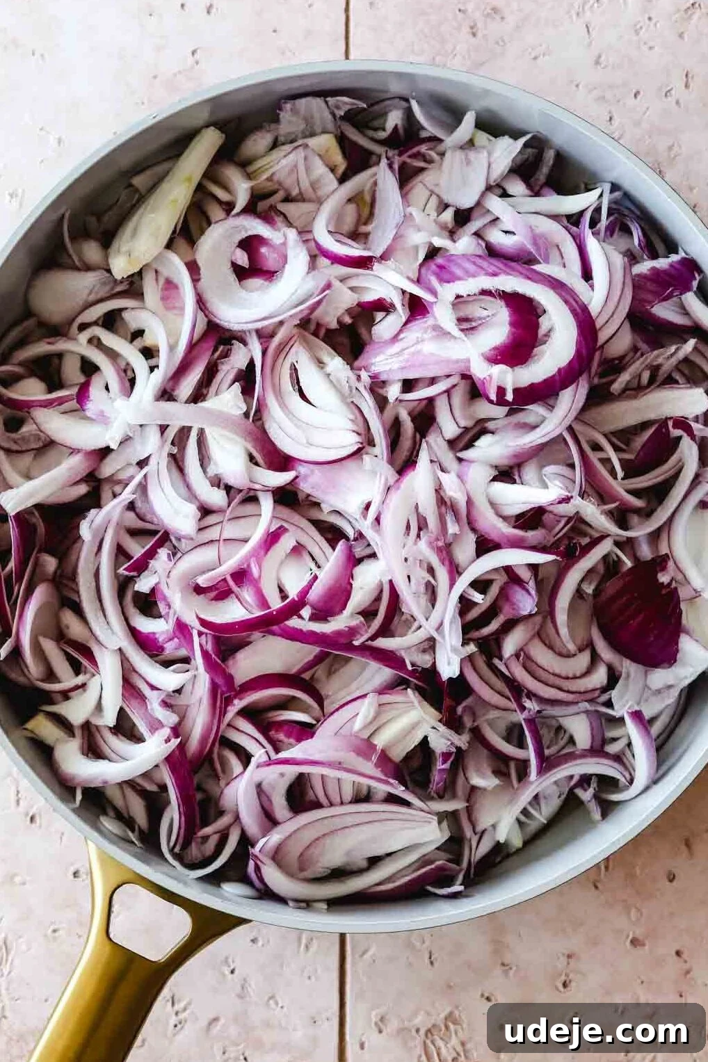 Sumac-Spiced Chicken Musakhan Crisps 10 Sliced red onions in a light coloured skillet with a gold handle on a light tiled background, prepared for caramelization.