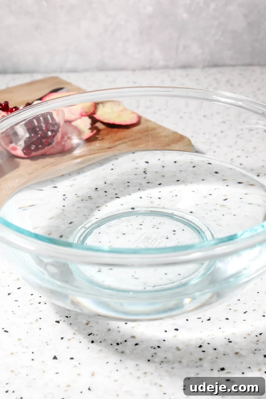 Homemade Pomegranate Molasses: Sweet, Tangy, and Versatile 12 large glass bowl with water halfway with cut up pomegranate on a wooden cutting board in the background on a speckled backdrop