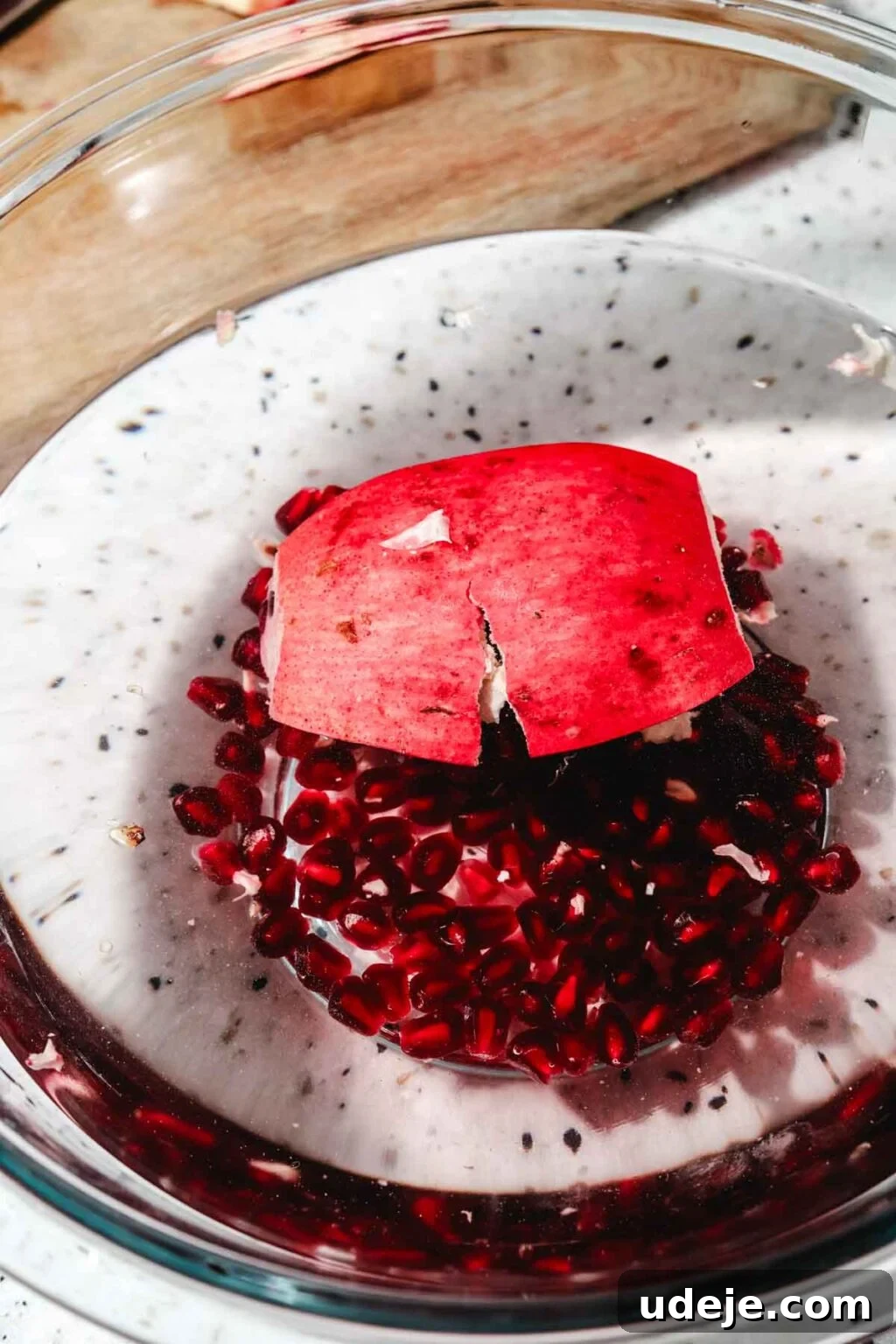 Homemade Pomegranate Molasses: Sweet, Tangy, and Versatile 13 piece of whole pomegranate in water with seeds floating in glass bowl with wooden cutting board at the back on a speckled background