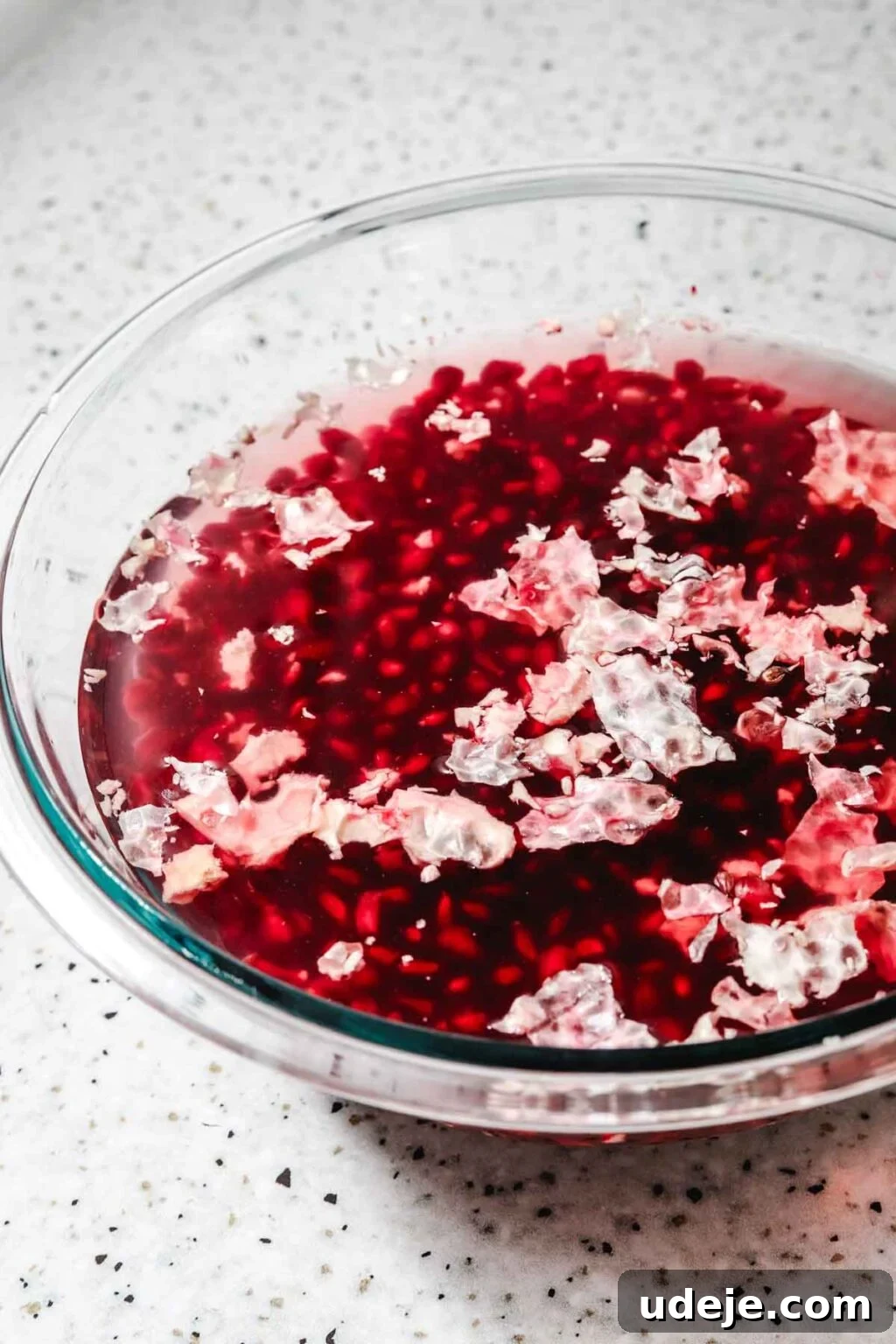 Homemade Pomegranate Molasses: Sweet, Tangy, and Versatile 14 pomegranate seeds in a glass bowl filled with water with the white pith floating to the top on a speckled background