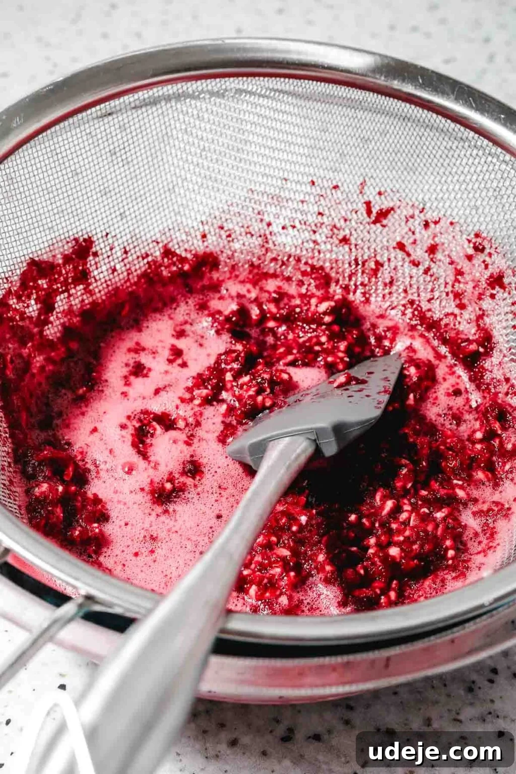 Homemade Pomegranate Molasses: Sweet, Tangy, and Versatile 19 blended pomegranate being sieved through a metal mesh sieve over a glass bowl with a grey spatula in it on a speckled background