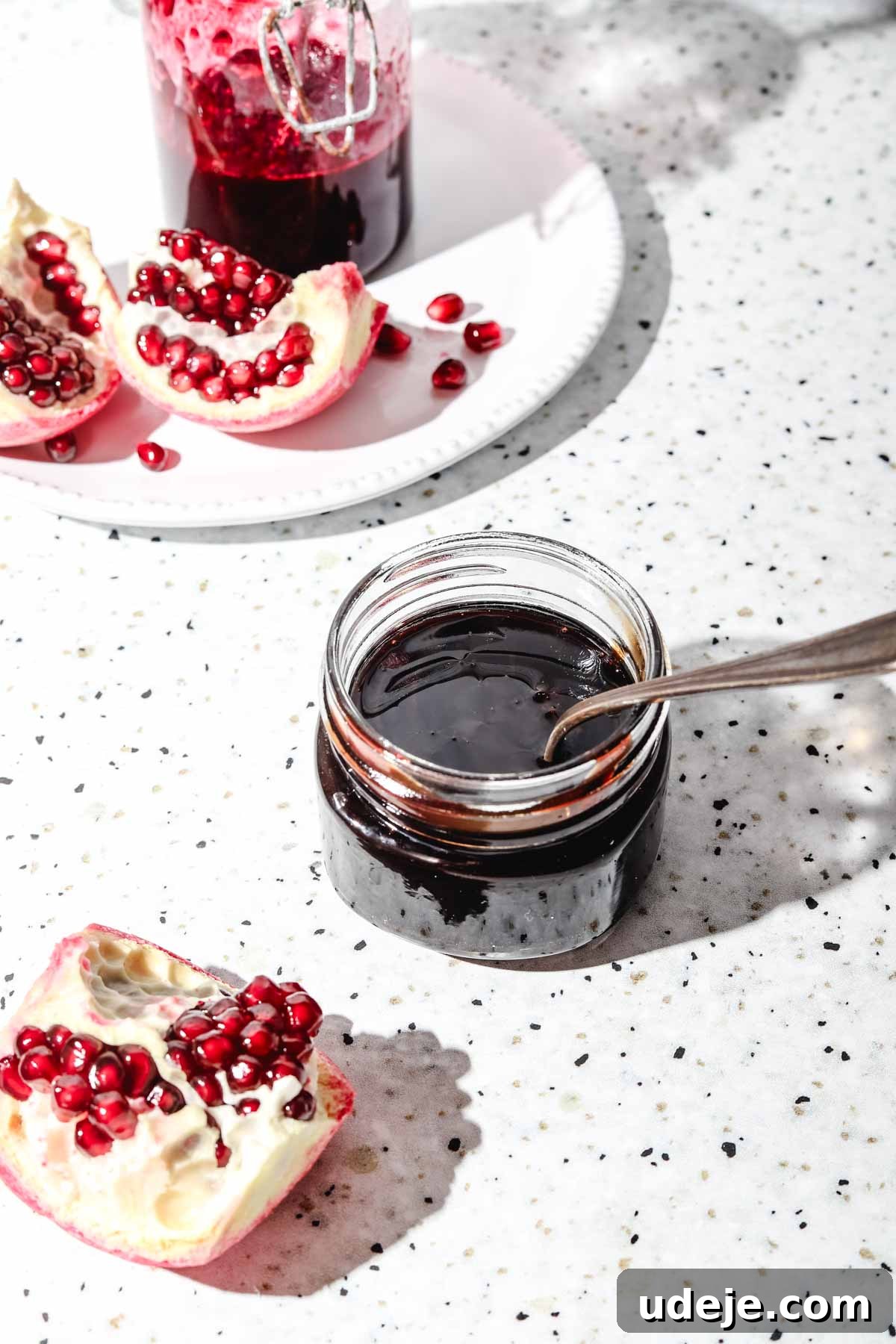 Homemade Pomegranate Molasses: Sweet, Tangy, and Versatile 3 Pomegranate molasses in a small mason jar with a spoon, alongside fresh pomegranate pieces on a white plate, set against a speckled background.