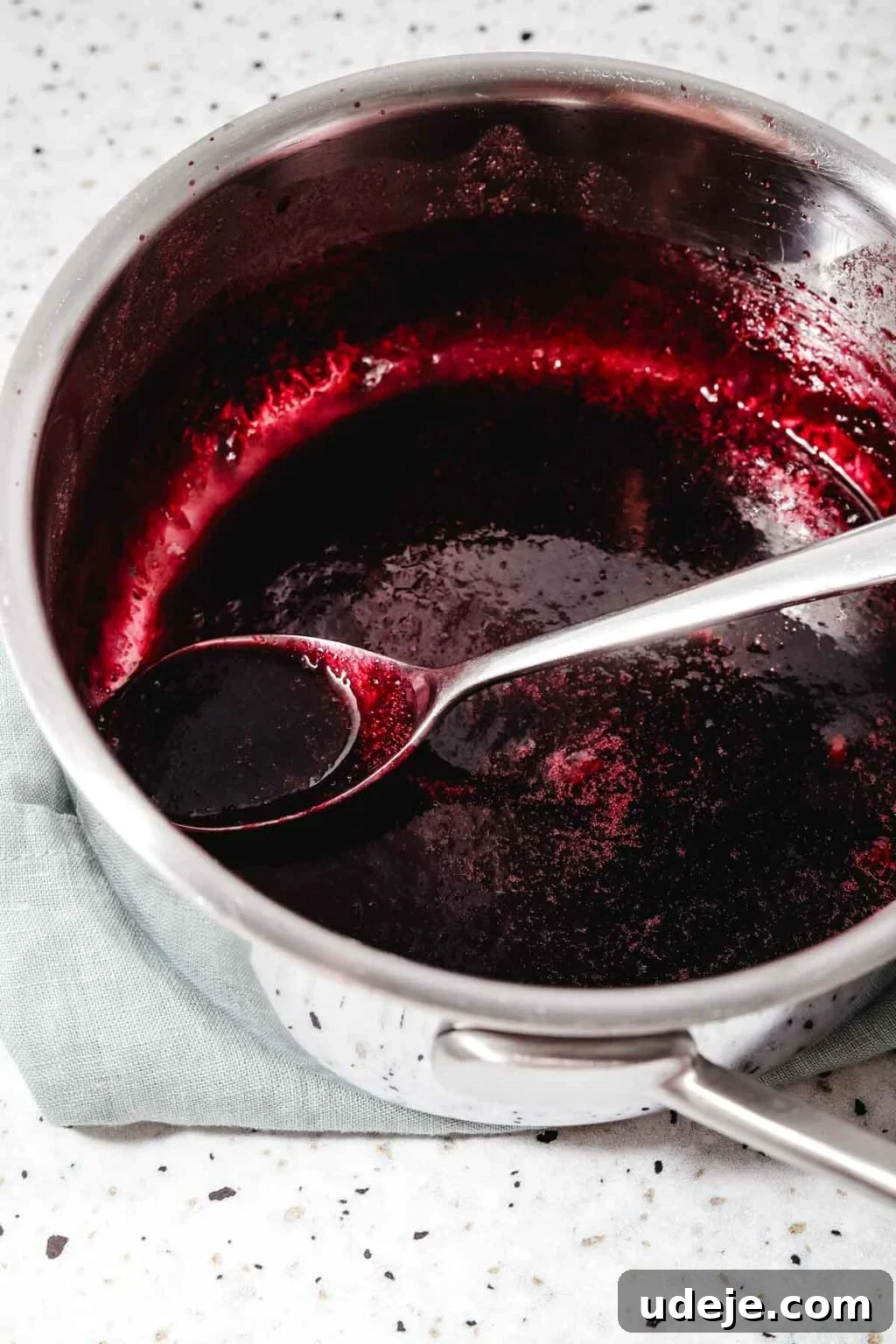 Homemade Pomegranate Molasses: Sweet, Tangy, and Versatile 22 reduced pomegranate juice in a metal saucepan with a metal spoon on a light green tea towel on a speckled background