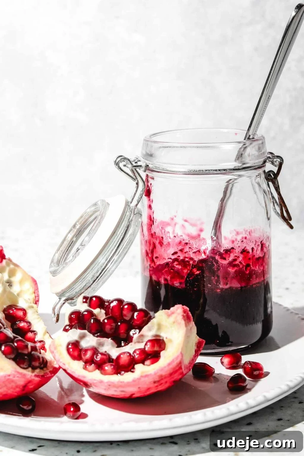 Homemade Pomegranate Molasses: Sweet, Tangy, and Versatile 23 pomegranate molasses in a small mason jar with a spoon in it with pieces of pomegranate beside it on a white plate on a speckled background