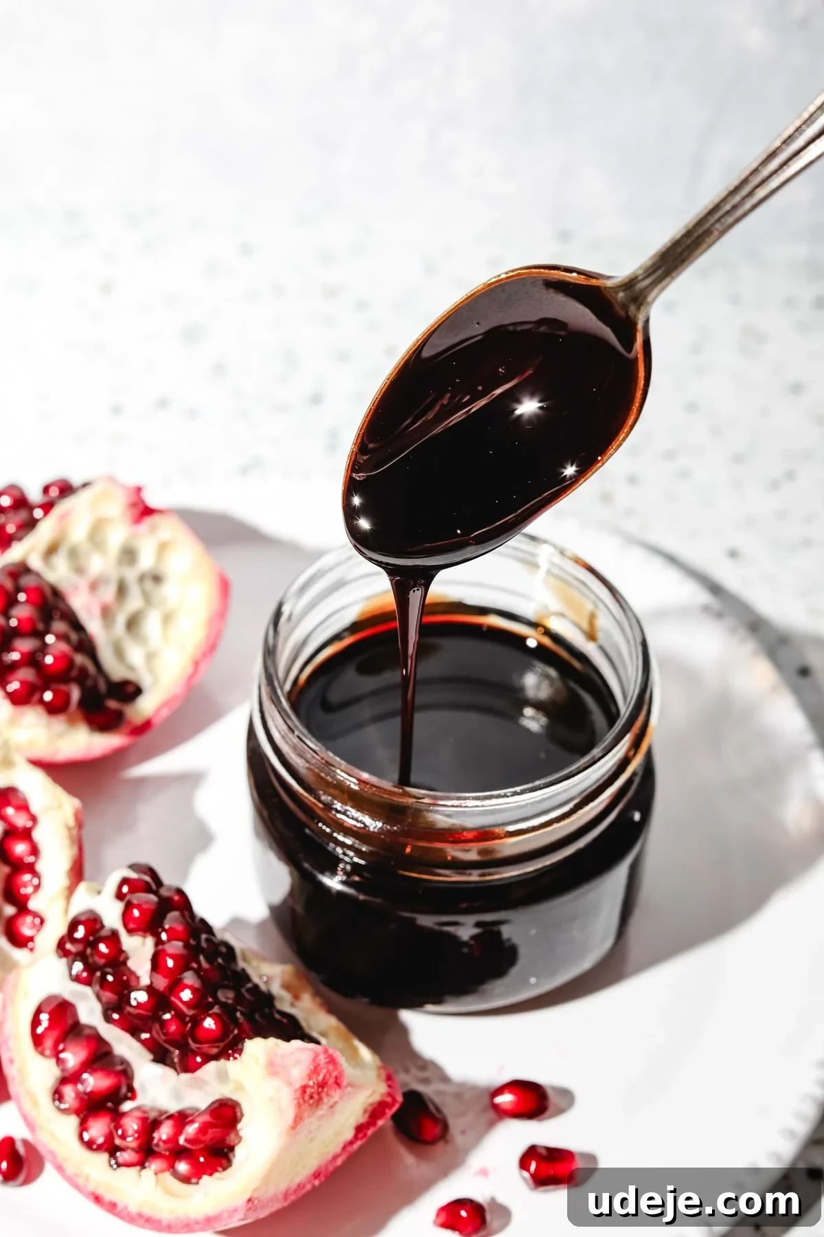 Homemade Pomegranate Molasses: Sweet, Tangy, and Versatile 4 Small jar of homemade pomegranate molasses with a spoon pouring it into the jar, placed on a white plate with a fresh pomegranate piece beside it on a speckled background.