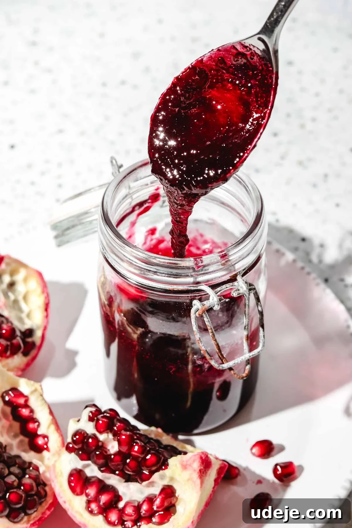 Homemade Pomegranate Molasses: Sweet, Tangy, and Versatile 5 Pomegranate molasses in a small mason jar with a spoon, alongside fresh pomegranate pieces on a white plate, set against a speckled background.